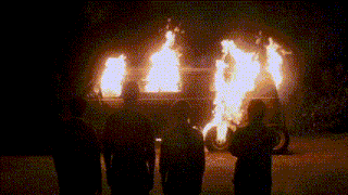 a group of people are standing in front of a burning van at night .