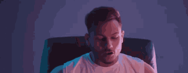 a man in a white tank top is sitting in a dark room with a purple light behind him .