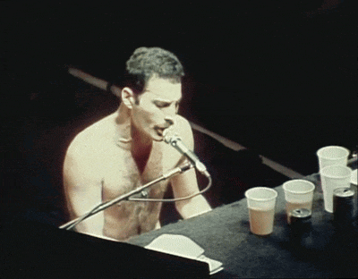 a shirtless man sings into a microphone while sitting at a keyboard