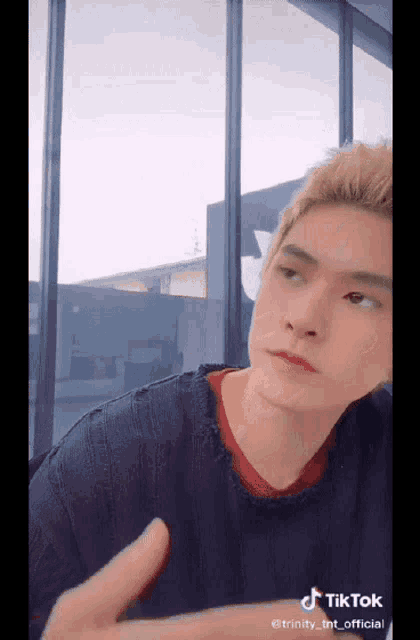 a man with blonde hair is sitting in front of a window and giving a thumbs up