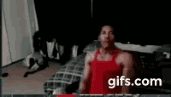 a man in a red tank top is standing in front of a bed in a dark room .