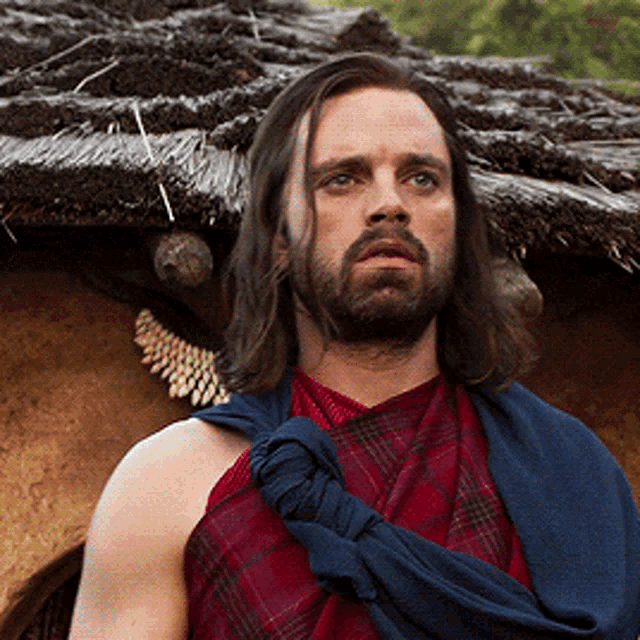 a man with long hair and a beard is standing in front of a thatched roof .