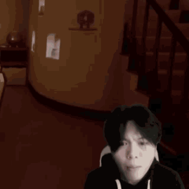 a man in a black hoodie is sitting in a dark room with a staircase in the background