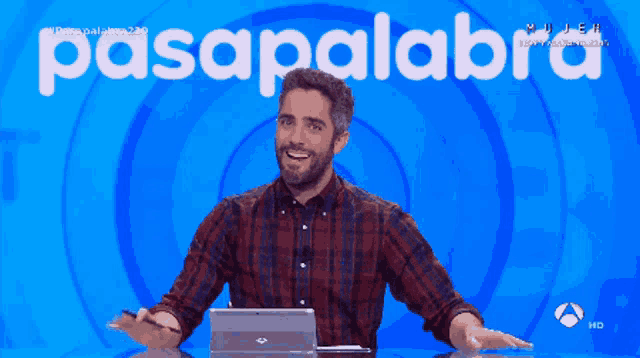 a man in a plaid shirt sits at a table with a laptop in front of a sign that says pasapalabra