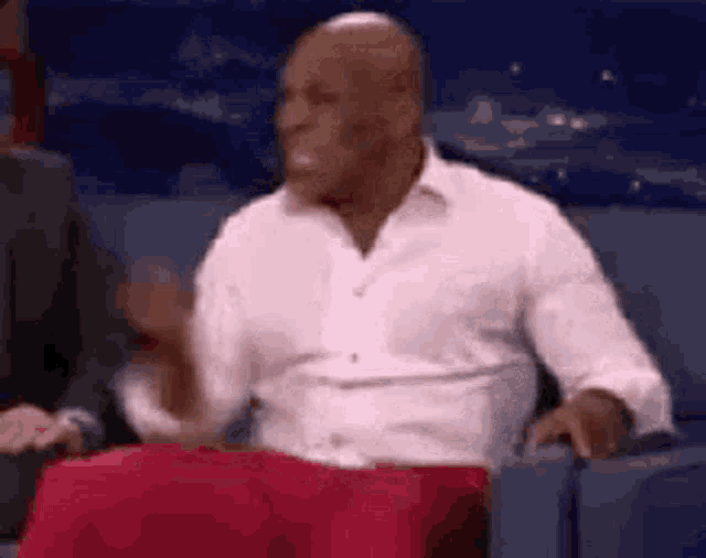 a man in a white shirt is sitting on a couch with a red pillow in front of him .