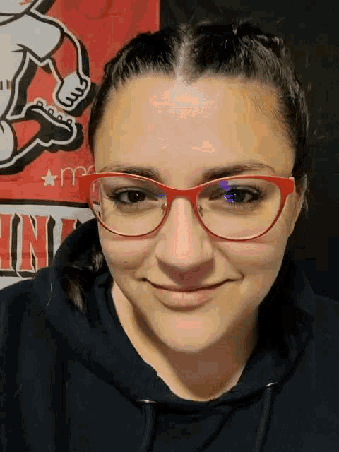 a woman wearing red glasses and a black hoodie smiles in front of a sign that says ' in '