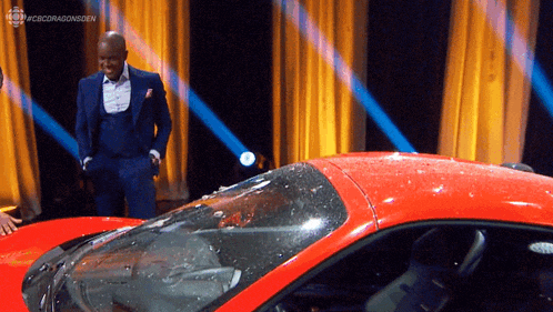 a man in a suit stands next to a red sports car on a stage with a cbs dragonsden logo on the bottom