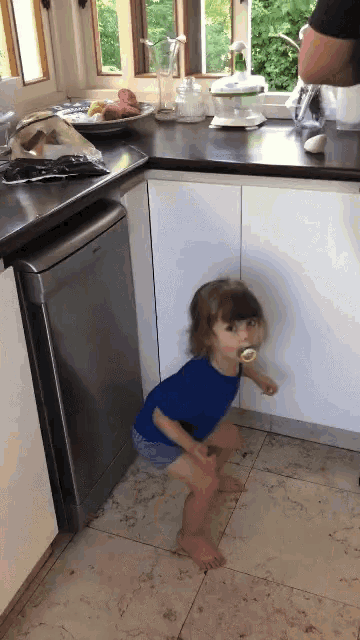 a little girl with a pacifier in her mouth is standing on the floor in a kitchen