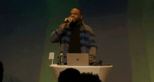 a man in a plaid shirt is standing at a podium with an apple laptop and a microphone .