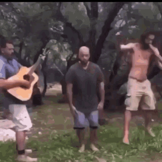 a man is playing a guitar while two men are dancing in the grass .
