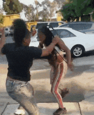 two women are fighting on a sidewalk in front of a parking lot .