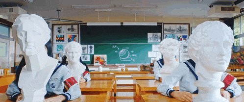 a group of students are sitting at desks in a classroom surrounded by busts of people