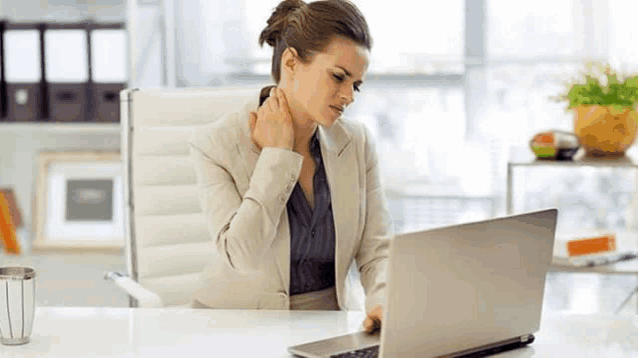 a woman is sitting at a desk in front of a laptop computer holding her neck .
