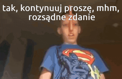a man in a superman shirt is standing in front of a sign that says tak kontynuuj prosze mhm rozsądne zanie
