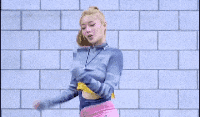 a woman in a blue sweater and pink shorts is dancing in front of a brick wall .
