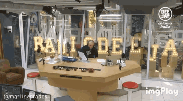 a man is sitting at a desk in front of a sign that says rai bella