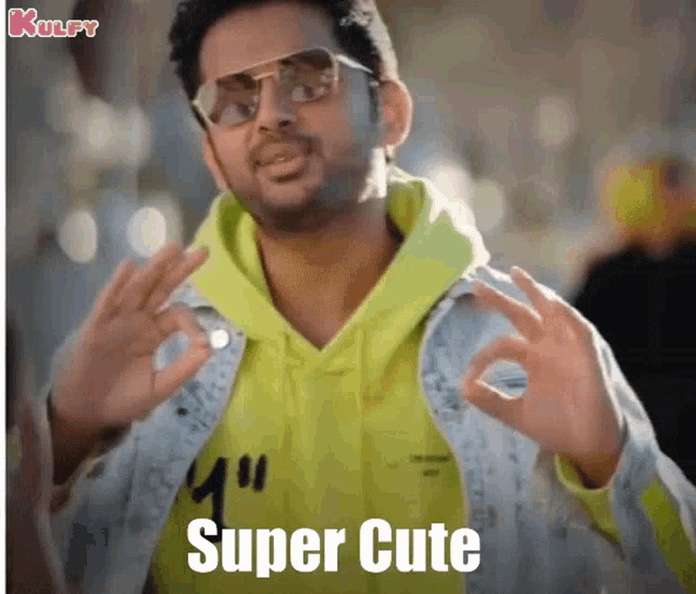 a man wearing sunglasses and a yellow hoodie is giving a super cute sign .