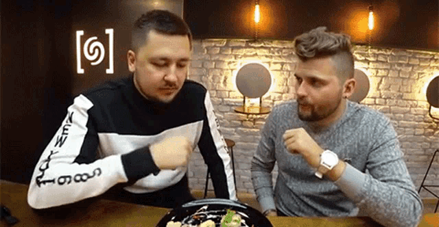 two men are sitting at a table with a plate of food and the letter s is on the wall
