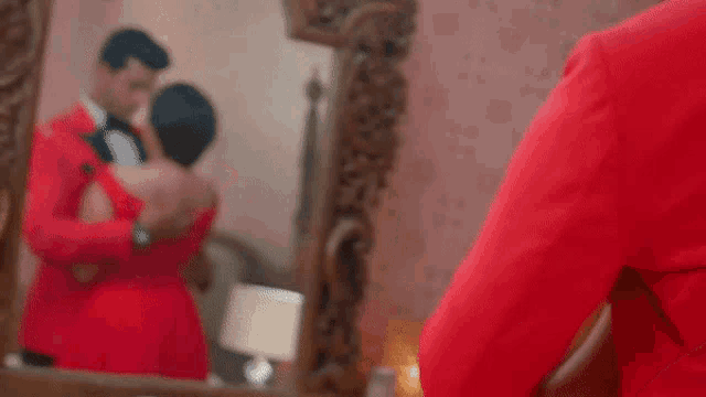 a man in a tuxedo is kissing a woman in a red dress in front of a mirror