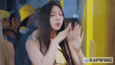 a woman in a yellow top is dancing in front of a yellow sign that says kapwing