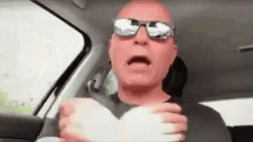 a bald man wearing sunglasses is driving a car and making a face .