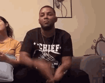 a man in a chief t-shirt is sitting on a couch with a woman behind him .