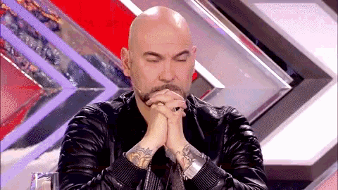 a bald man with a beard is sitting with his hands folded in prayer