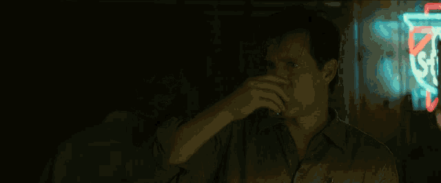 a man in a green shirt is smoking a cigarette in front of a neon sign that says ' s ' on it