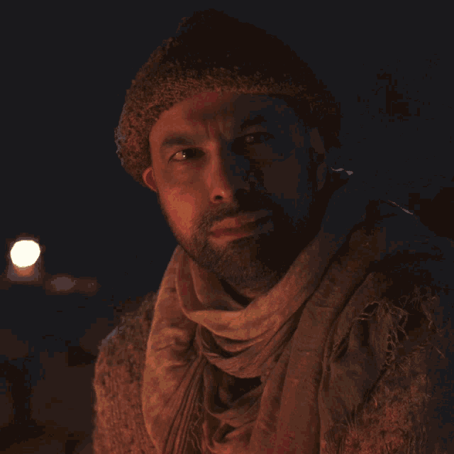 a man with a beard wearing a beanie and scarf looks down at something