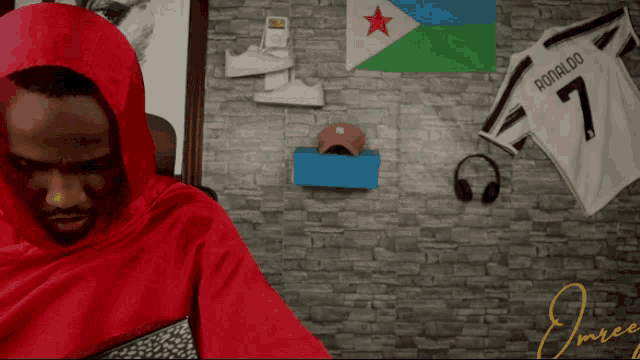 a man in a red hoodie is sitting in front of a brick wall with a ronaldo 7 jersey hanging on it
