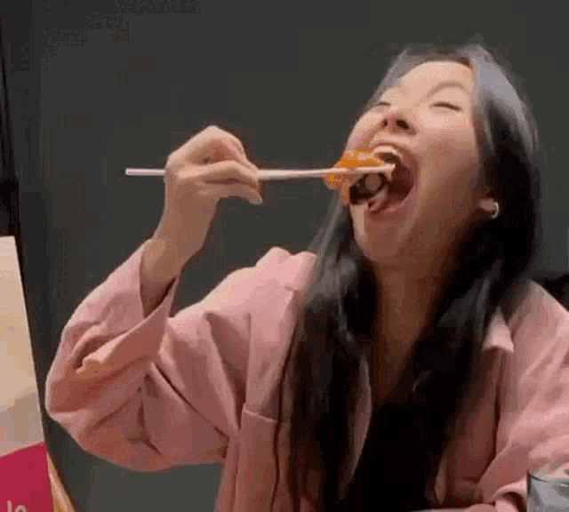 a woman in a pink jacket is eating food with chopsticks and making a funny face .