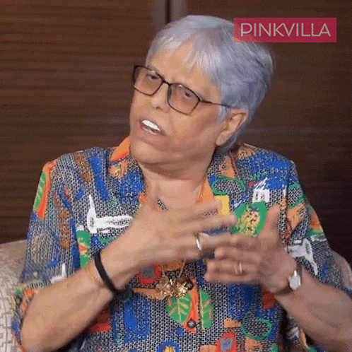 an older woman wearing glasses and a colorful shirt is sitting on a couch with a pinkvilla sign above her