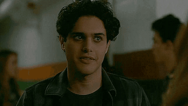 a young man with curly hair is wearing a black jacket and a black shirt and looking at the camera .