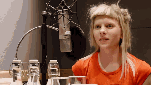 a woman in an orange shirt stands in front of a microphone with a sticker on it that says ' a '