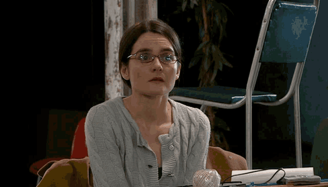 a woman wearing glasses and a grey sweater sits at a desk