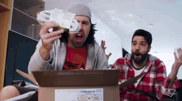 two men are sitting in front of a cardboard box . one of the men is holding a remote control .