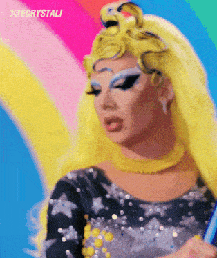 a close up of a drag queen 's face with a rainbow in the background and the words xtecrystali above her