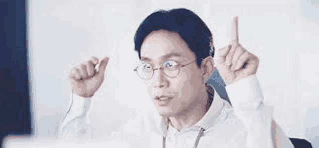 a man wearing glasses is pointing up while sitting in front of a computer screen .