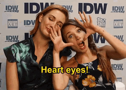two women making faces in front of a wall that says heart eyes