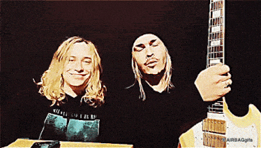 a man holding a guitar next to another man with long hair