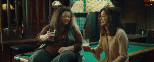 two women are sitting at a pool table with one holding a glass of beer