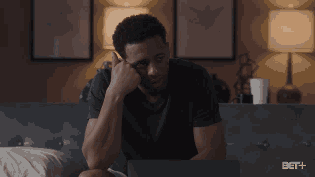 a man sits on a bed looking at a laptop screen that says bet + on the bottom