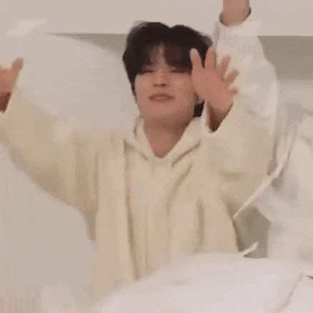 a man in a white hoodie is sitting on a bed with his arms in the air .