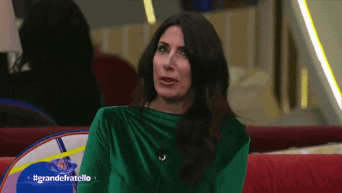 a woman in a green dress is sitting on a red couch in front of a sign that says grandefratello on it
