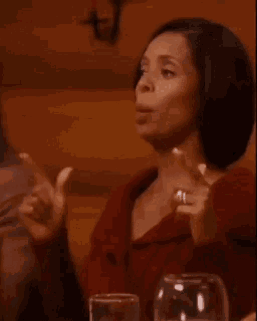a woman in a red dress is sitting at a table with a glass of water and pointing at something .