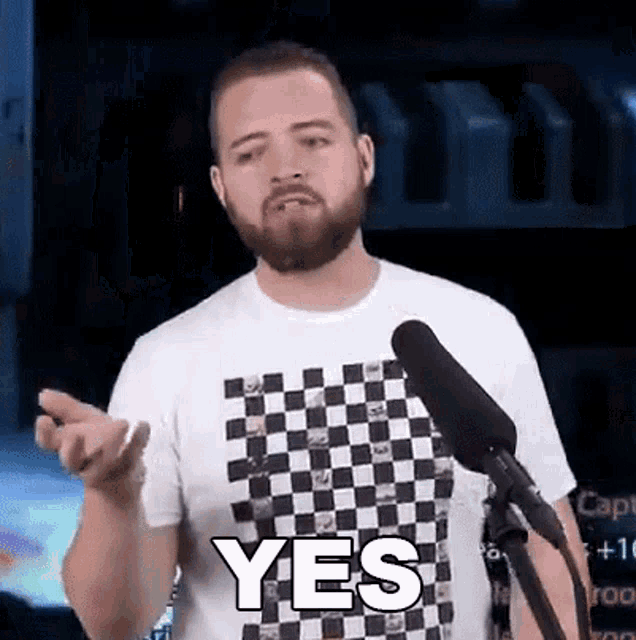 a man with a beard and a checkered shirt is standing in front of a microphone .