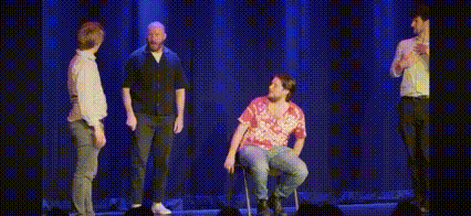 a group of men are standing on a stage and one of them is sitting in a chair and saying `` i hate you '' .