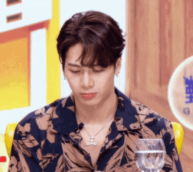 a young man wearing a necklace and a floral shirt is sitting next to a glass of water