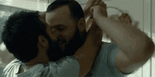 a man with a beard is kissing another man on the cheek in a kitchen .