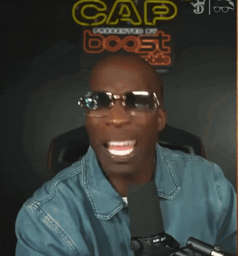 a man wearing sunglasses and a blue shirt is laughing in front of a sign that says cap